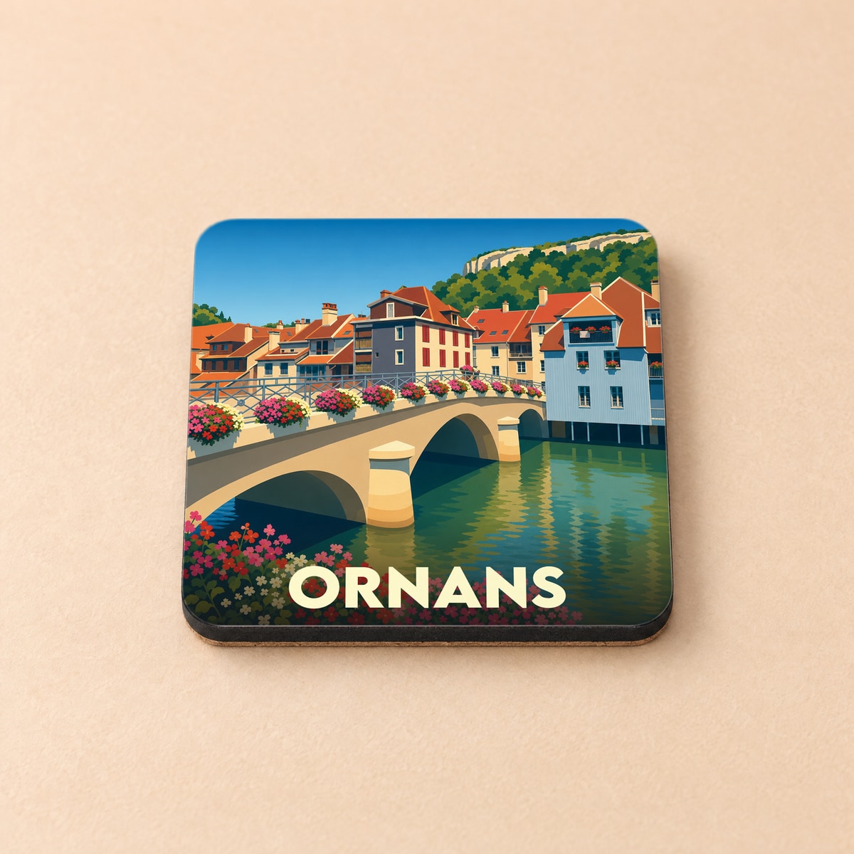 Dessous de verre souvenir de la ville d'Ornans avec une illustration stylisée d'un pont fleuri et de maisons colorées au bord de l'eau.