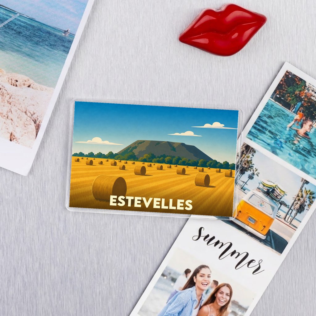 Magnet souvenir de la ville d'Estevelles avec une illustration stylisée d'un champ de meules de foin devant une montagne.