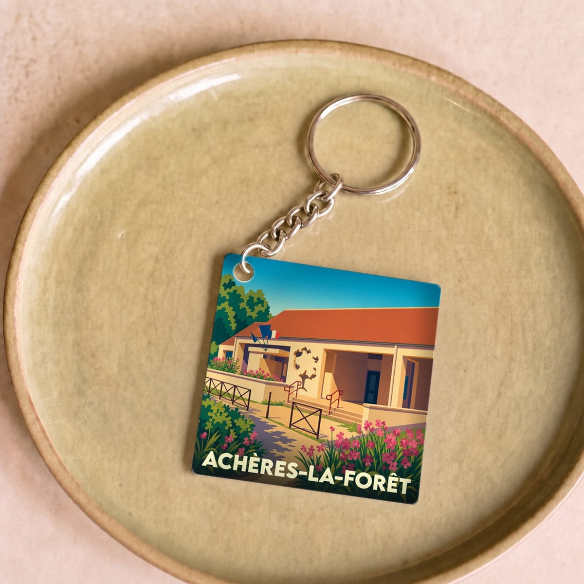 Porte-clés souvenir de la ville d'Achères-la-Forêt avec une illustration stylisée d'un bâtiment fleuri.