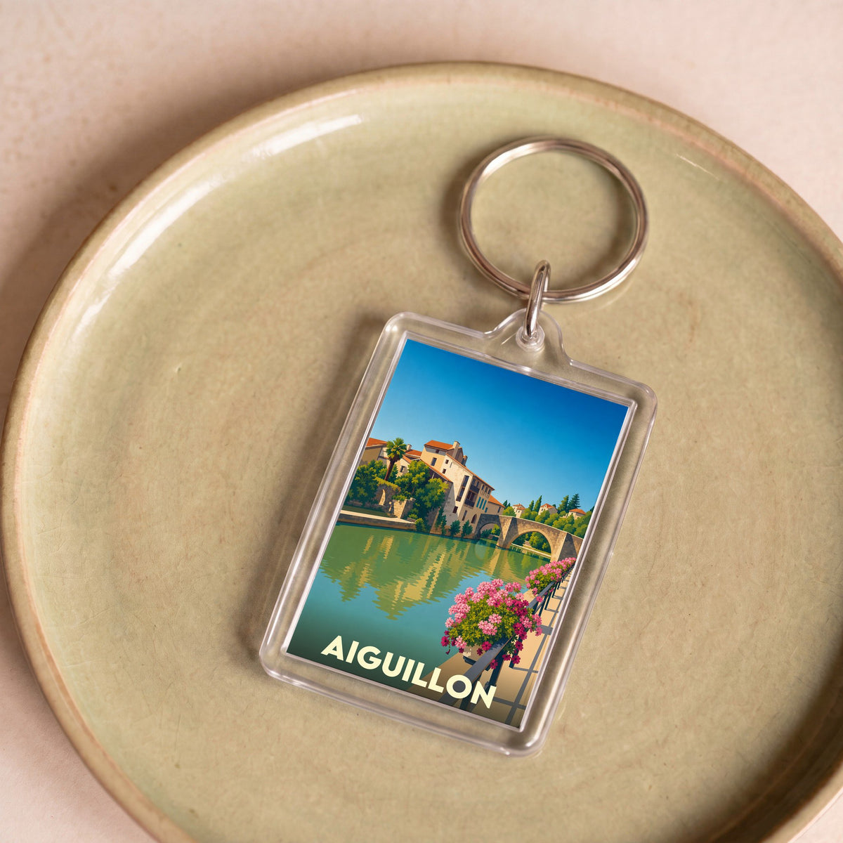 Porte-clés souvenir de la ville d'Aiguillon avec une illustration stylisée d'un pont et de maisons au bord de l'eau.