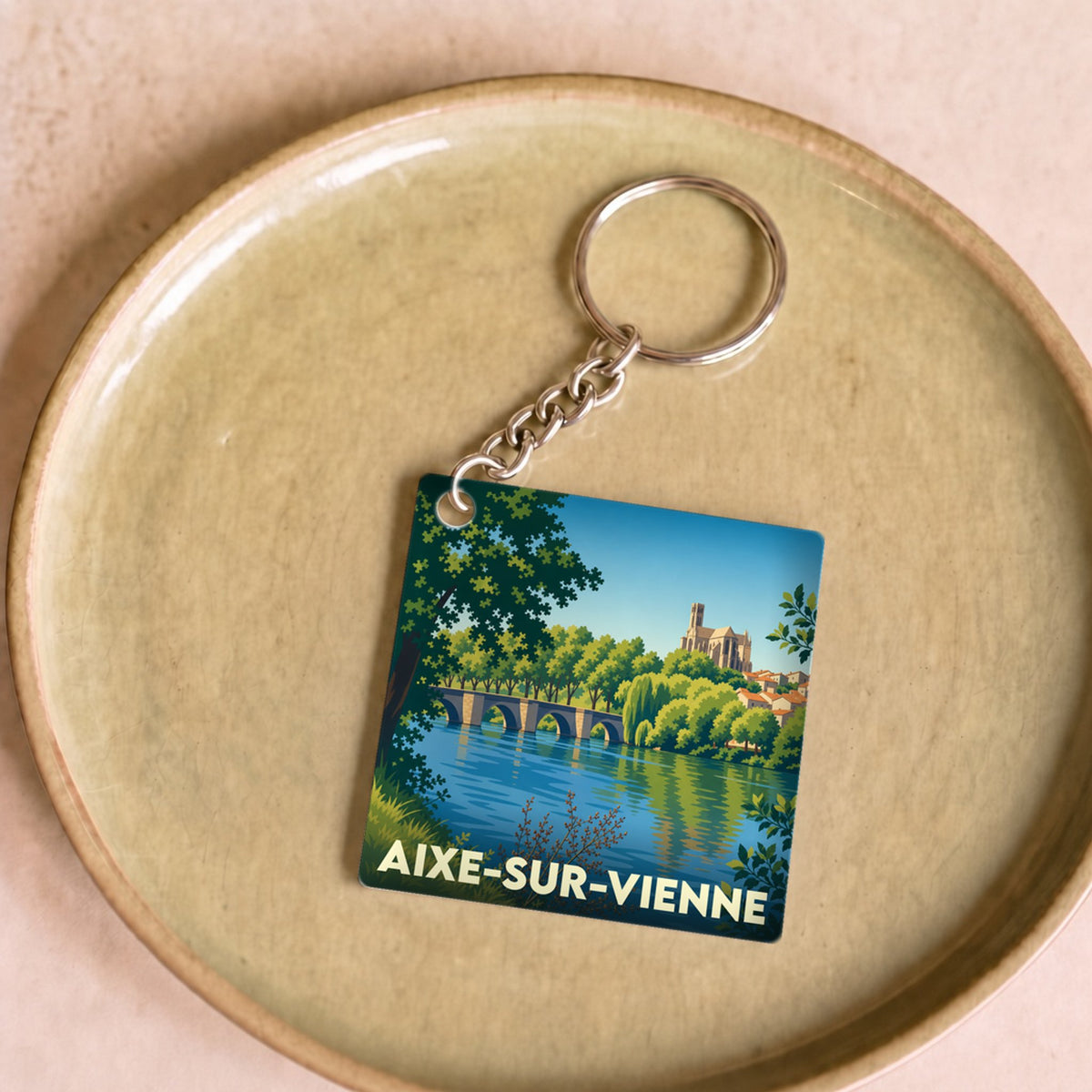 Porte-clés souvenir de la ville d'Aixe-sur-Vienne avec une illustration stylisée d'un pont et d'une église au bord de l'eau.