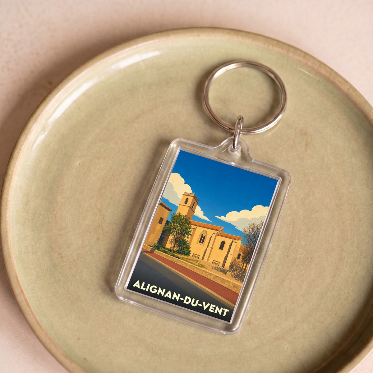 Porte-clés souvenir de la ville d'Alignan-du-Vent avec une illustration stylisée de son église sous un ciel bleu.