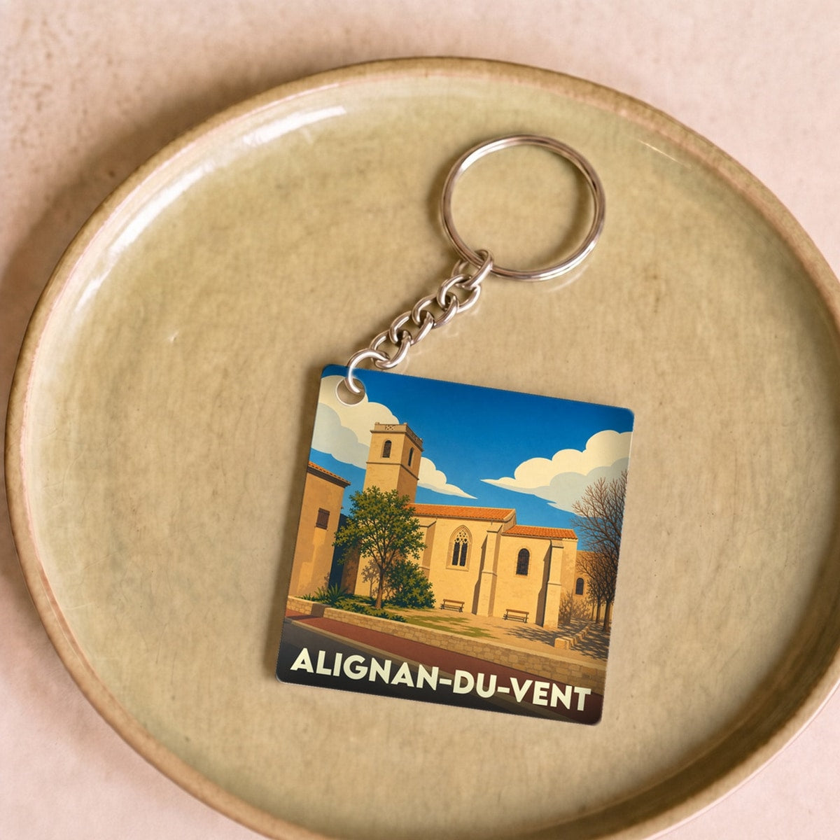Porte-clés souvenir de la ville d'Alignan-du-Vent avec une illustration stylisée de son église sous un ciel bleu.