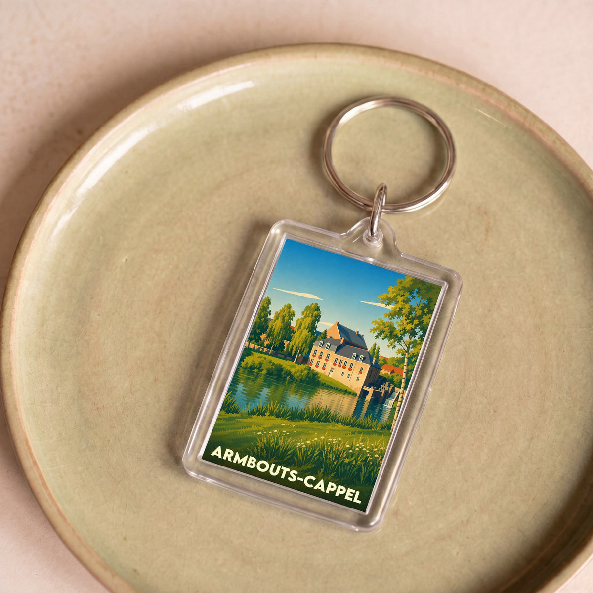 Porte-clés souvenir de la ville d'Armbouts-Cappel avec une illustration stylisée d'un paysage champêtre au bord de l'eau.