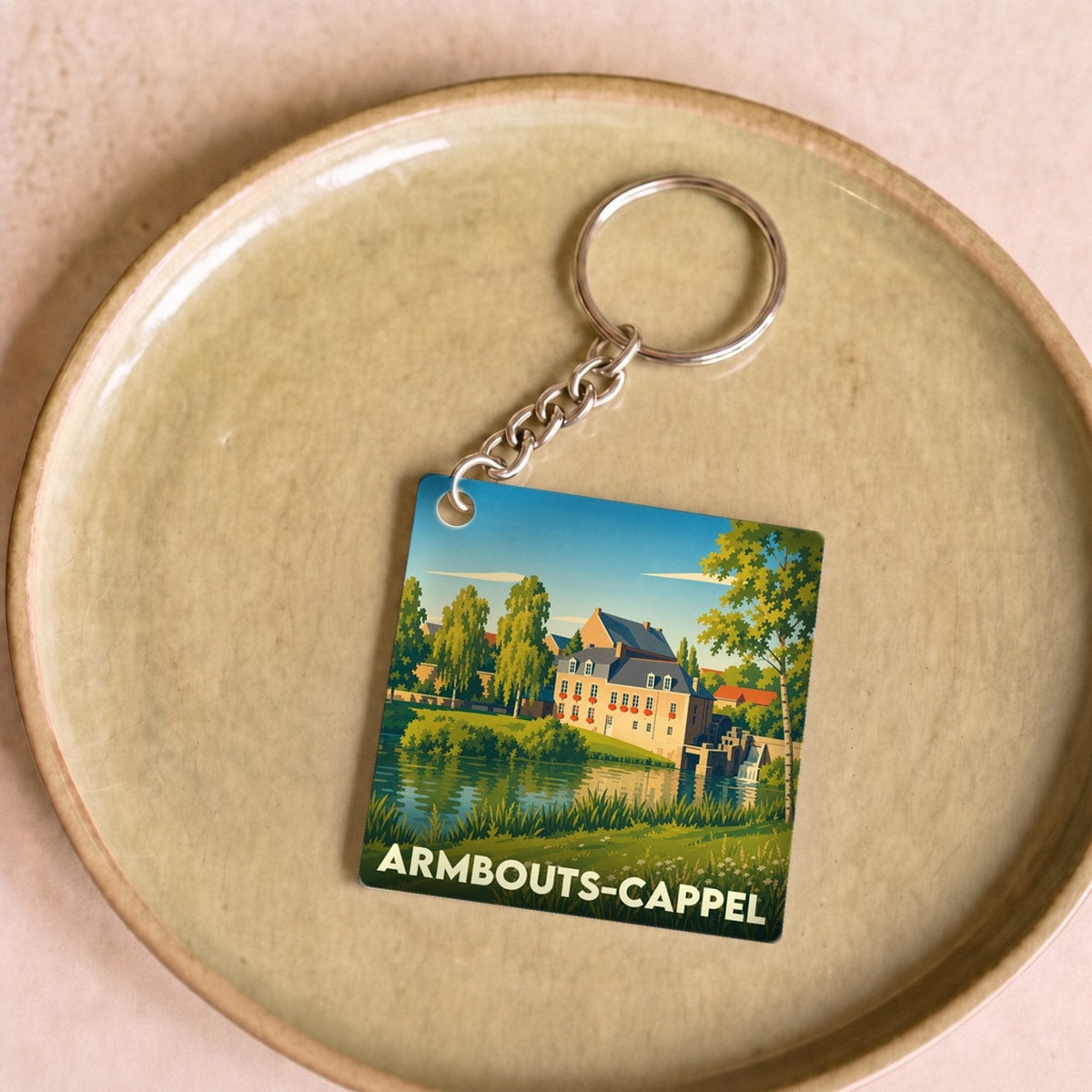 Porte-clés souvenir de la ville d'Armbouts-Cappel avec une illustration stylisée d'un paysage champêtre au bord de l'eau.