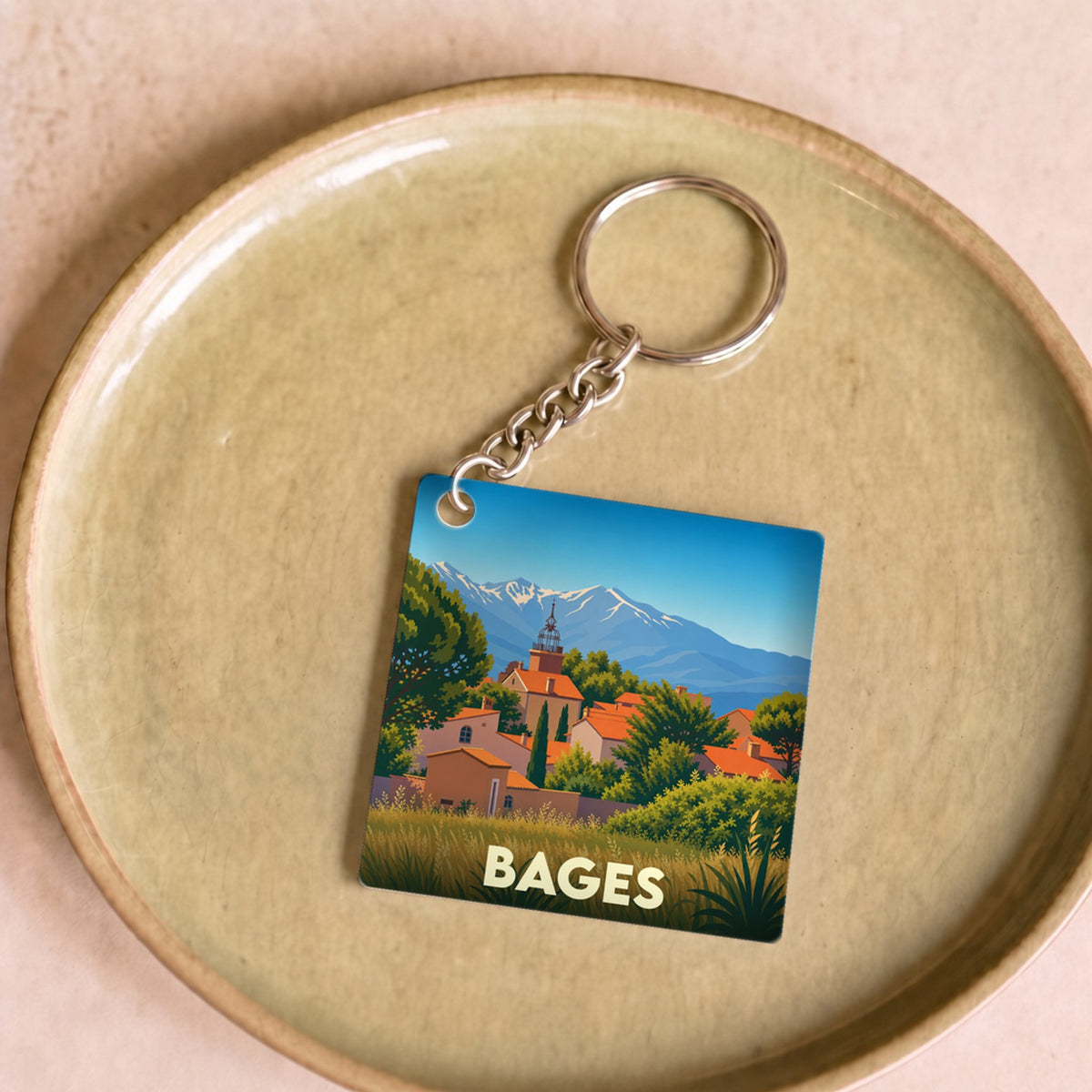 Porte-clés souvenir de la ville de Bages avec une illustration stylisée d'un village face aux montagnes.