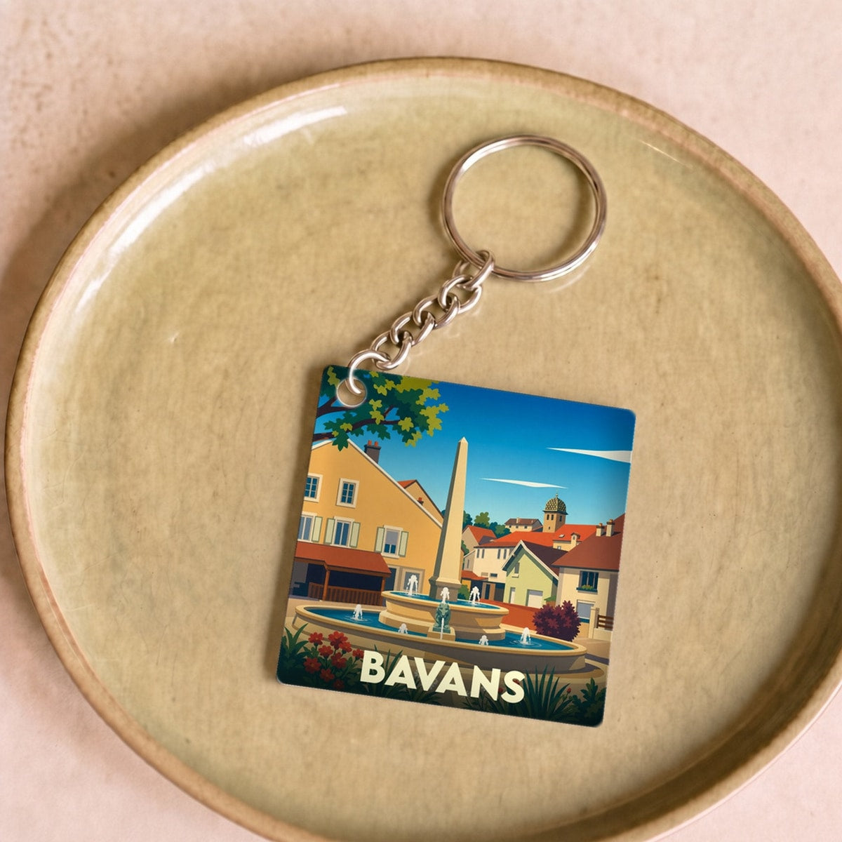 Porte-clés souvenir de la ville de Bavans illustrant une place ensoleillée avec une fontaine.