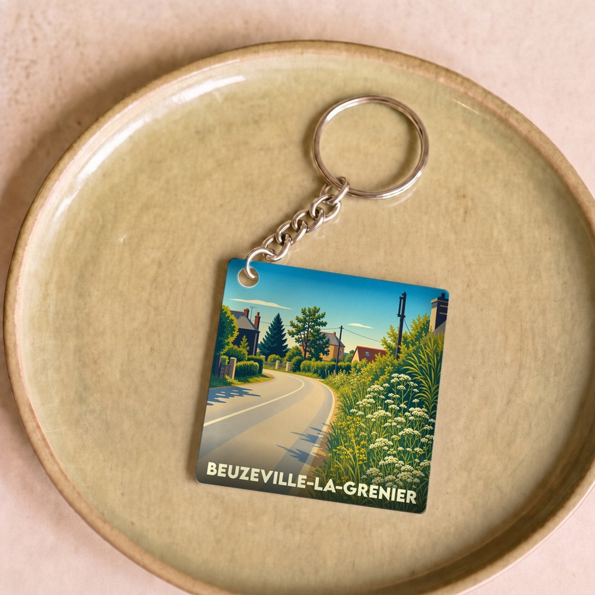 Porte-clés souvenir de Beuzeville-la-Grenier avec une illustration stylisée de route de campagne fleurie.
