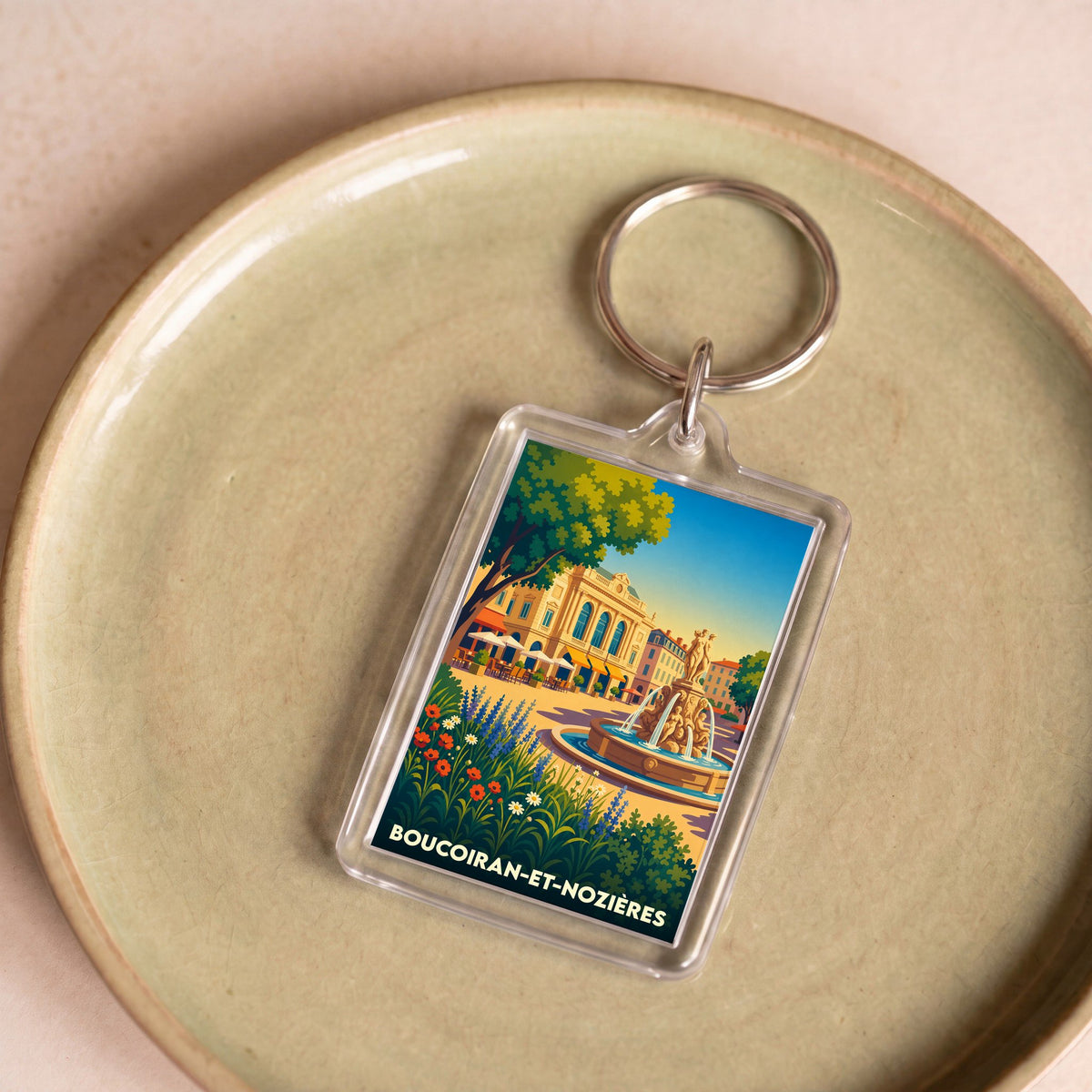 Porte-clés souvenir de la ville de Boucoiran-et-Nozières avec une illustration colorée et stylisée d'une place ensoleillée.