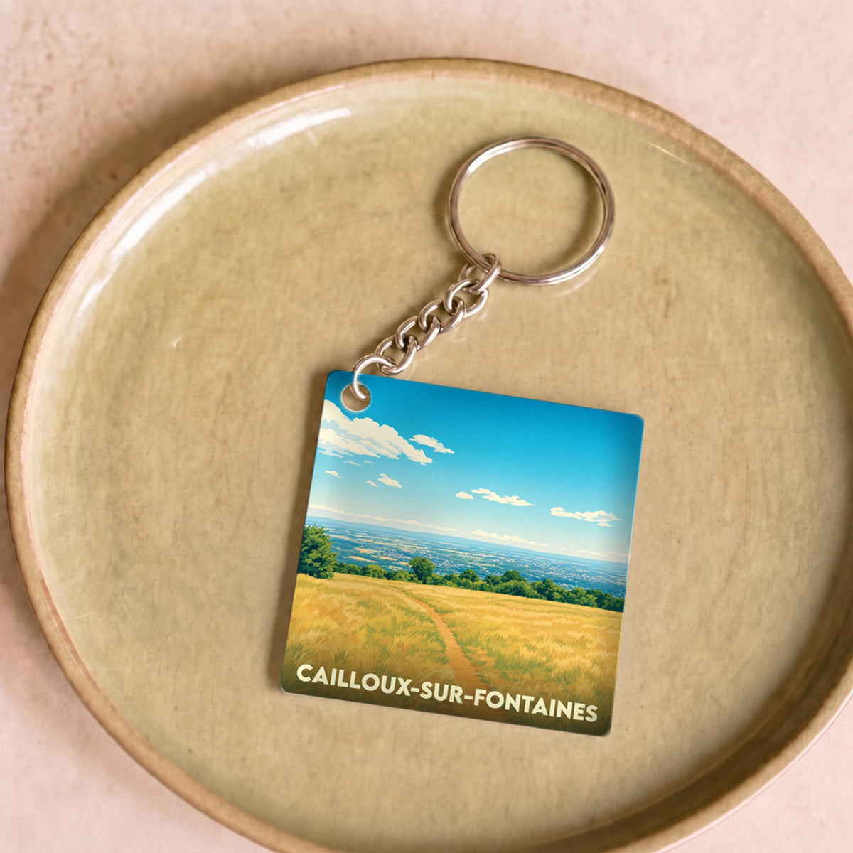 Porte-clés souvenir de la ville de Cailloux-sur-Fontaines avec une illustration stylisée d'un paysage champêtre.