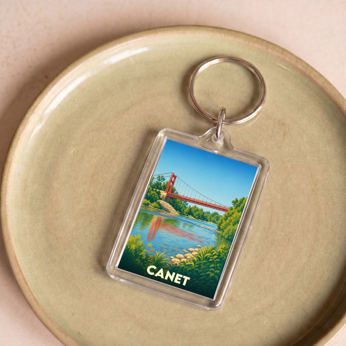 Porte-clés souvenir de la ville de Canet avec une illustration stylisée d'un pont suspendu rouge sur une rivière.