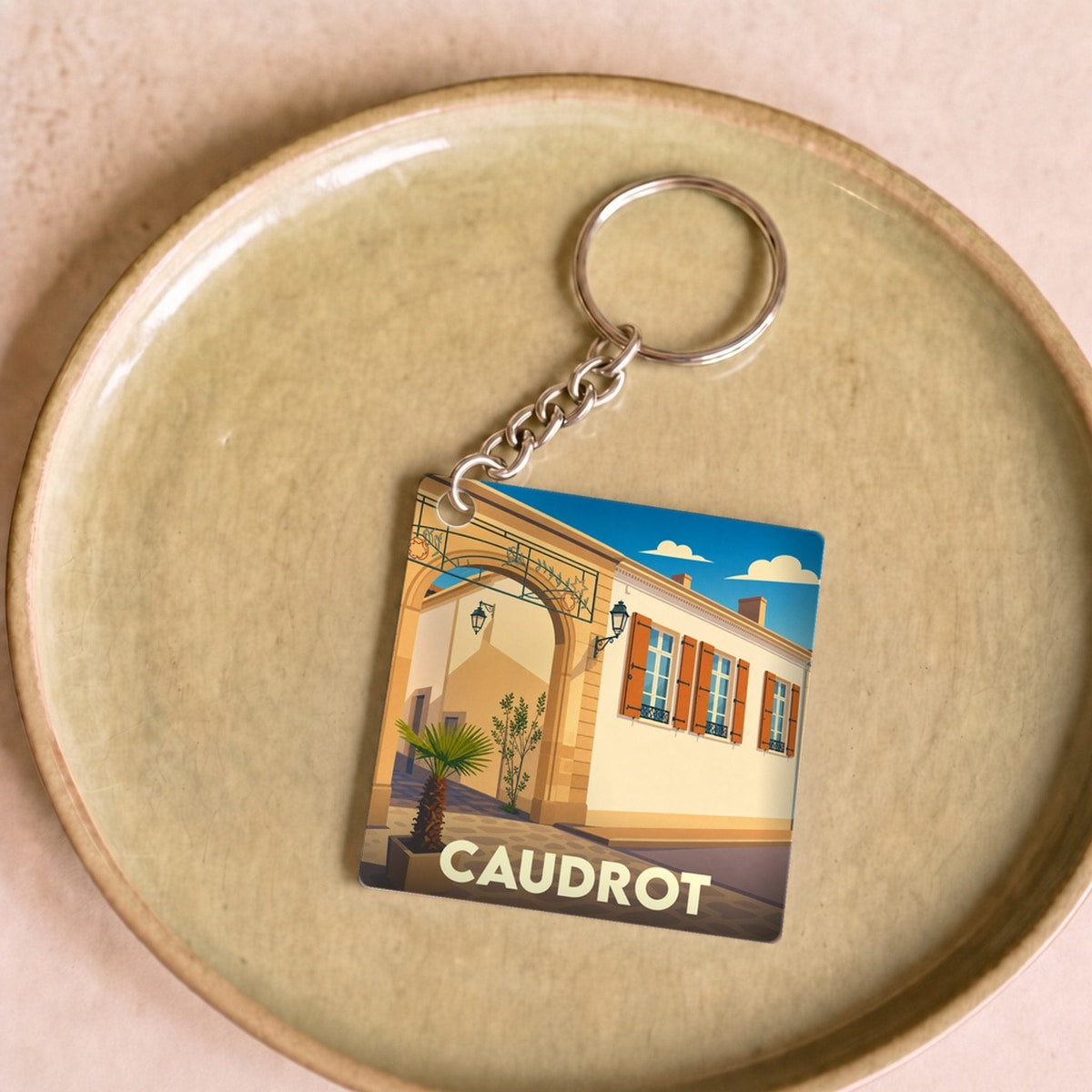 Porte-clés souvenir de la ville de Caudrot avec une illustration stylisée d'une rue ensoleillée.