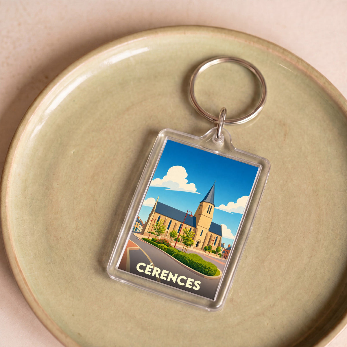 Porte-clés souvenir de la ville de Cérences avec une illustration stylisée de son église sous un ciel bleu.
