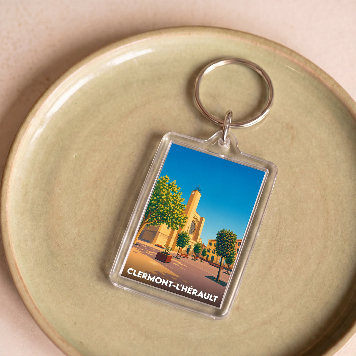 Porte-clés souvenir de la ville de Clermont-l'Hérault avec une illustration stylisée de son église et de ses arbres.