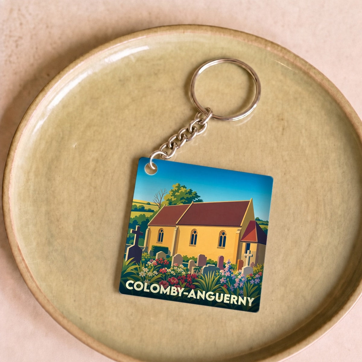 Porte-clés souvenir de la ville de Colomby-Anguerny avec une illustration stylisée de son église et de son cimetière fleuri.