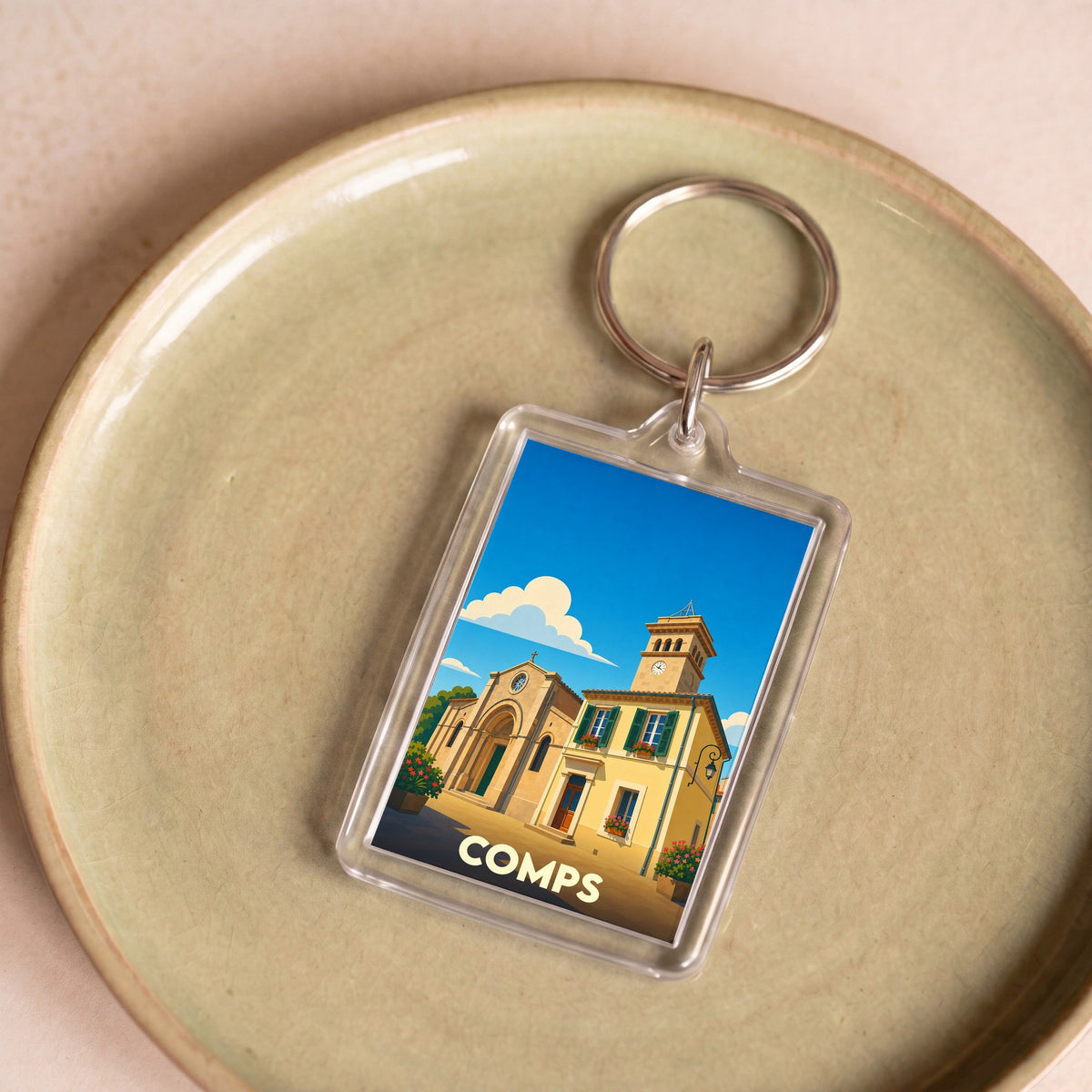 Porte-clés souvenir de la ville de Comps avec une illustration stylisée de son église sous un ciel bleu.