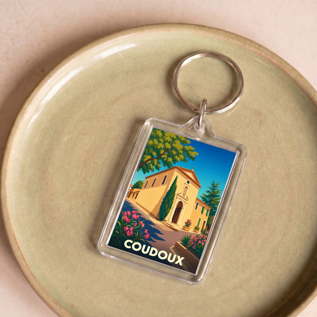 Porte-clés souvenir de la ville de Coudoux avec une illustration stylisée de son église provençale sous un ciel bleu.