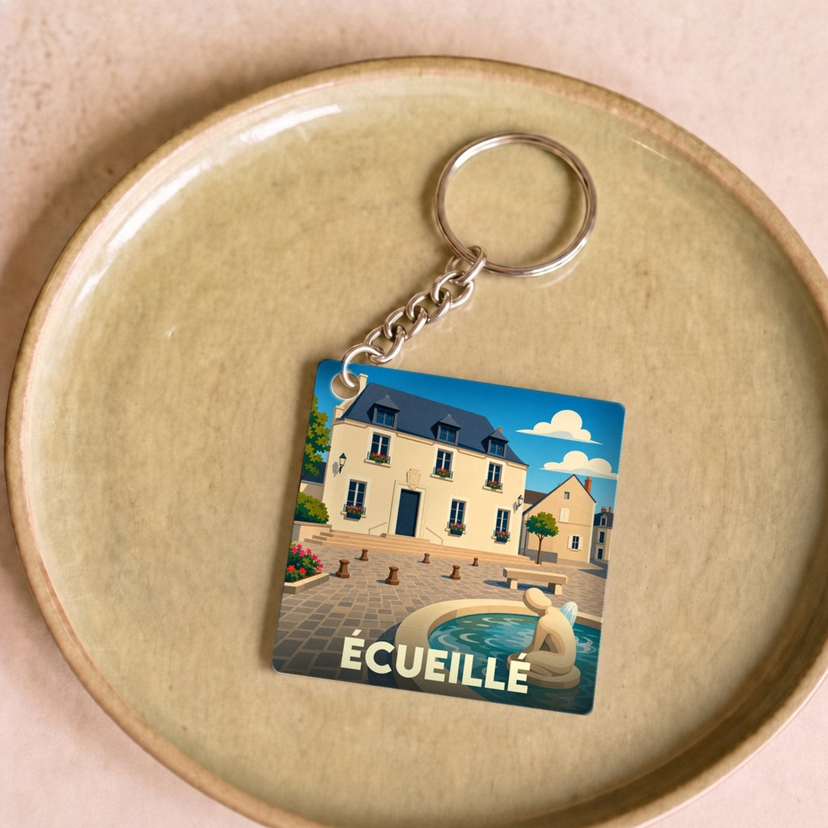 Porte-clés souvenir de la ville d’Écueillé avec une illustration stylisée d'une place et sa fontaine.