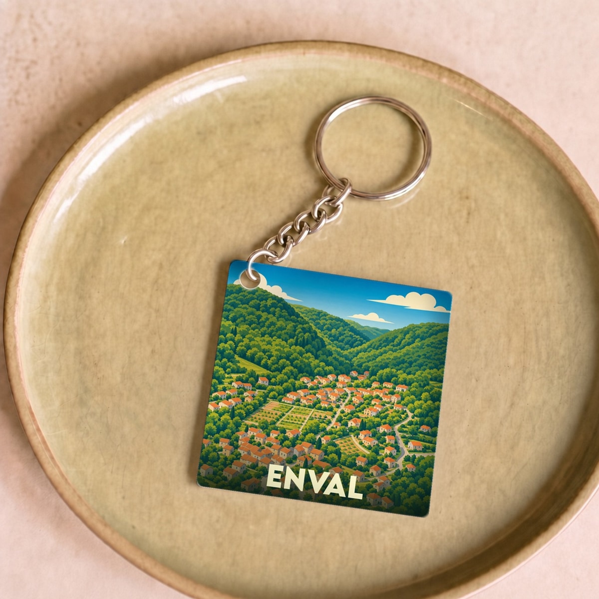 Porte-clés souvenir de la ville d'Enval avec une illustration stylisée d'un village dans une vallée verdoyante.