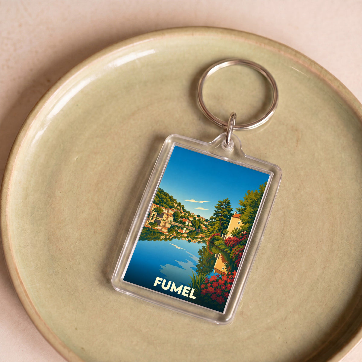 Porte-clés souvenir de la ville de Fumel avec une illustration stylisée représentant un paysage au bord de l'eau.