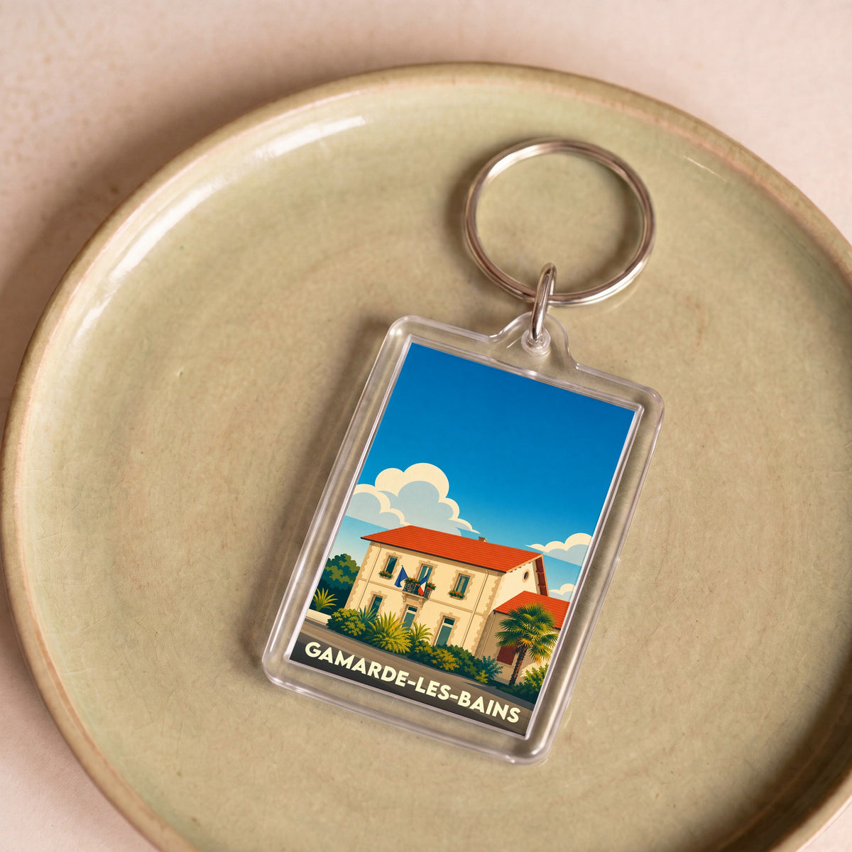 Porte-clés souvenir de la ville de Gamarde-les-Bains avec une illustration stylisée d'une maison landaise sous un ciel bleu.