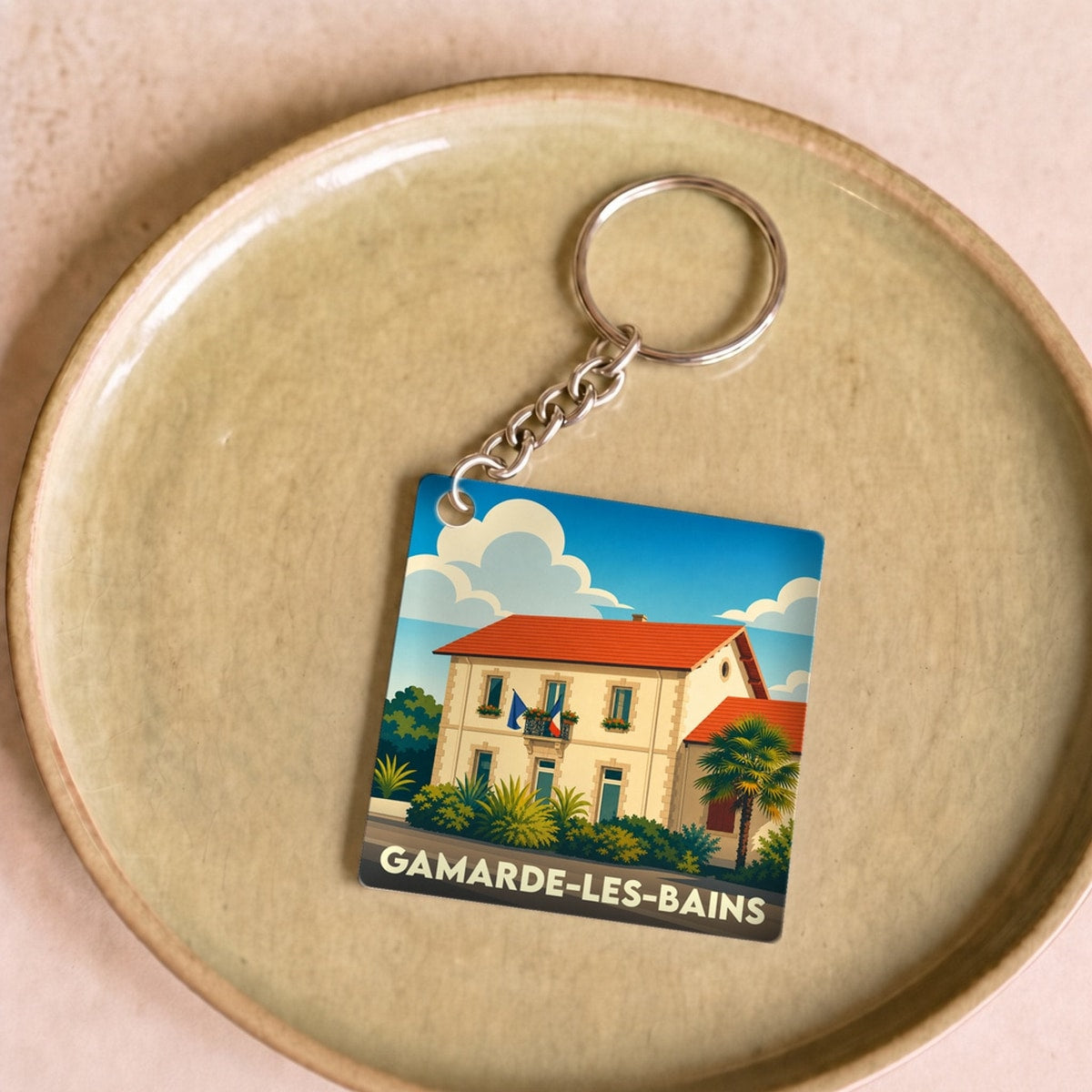 Porte-clés souvenir de la ville de Gamarde-les-Bains avec une illustration stylisée d'une maison landaise sous un ciel bleu.