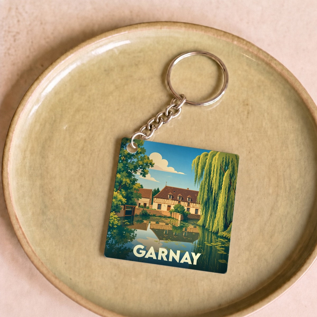Porte-clés souvenir de la ville de Garnay avec une illustration stylisée d'un paysage bucolique au bord de l'eau.