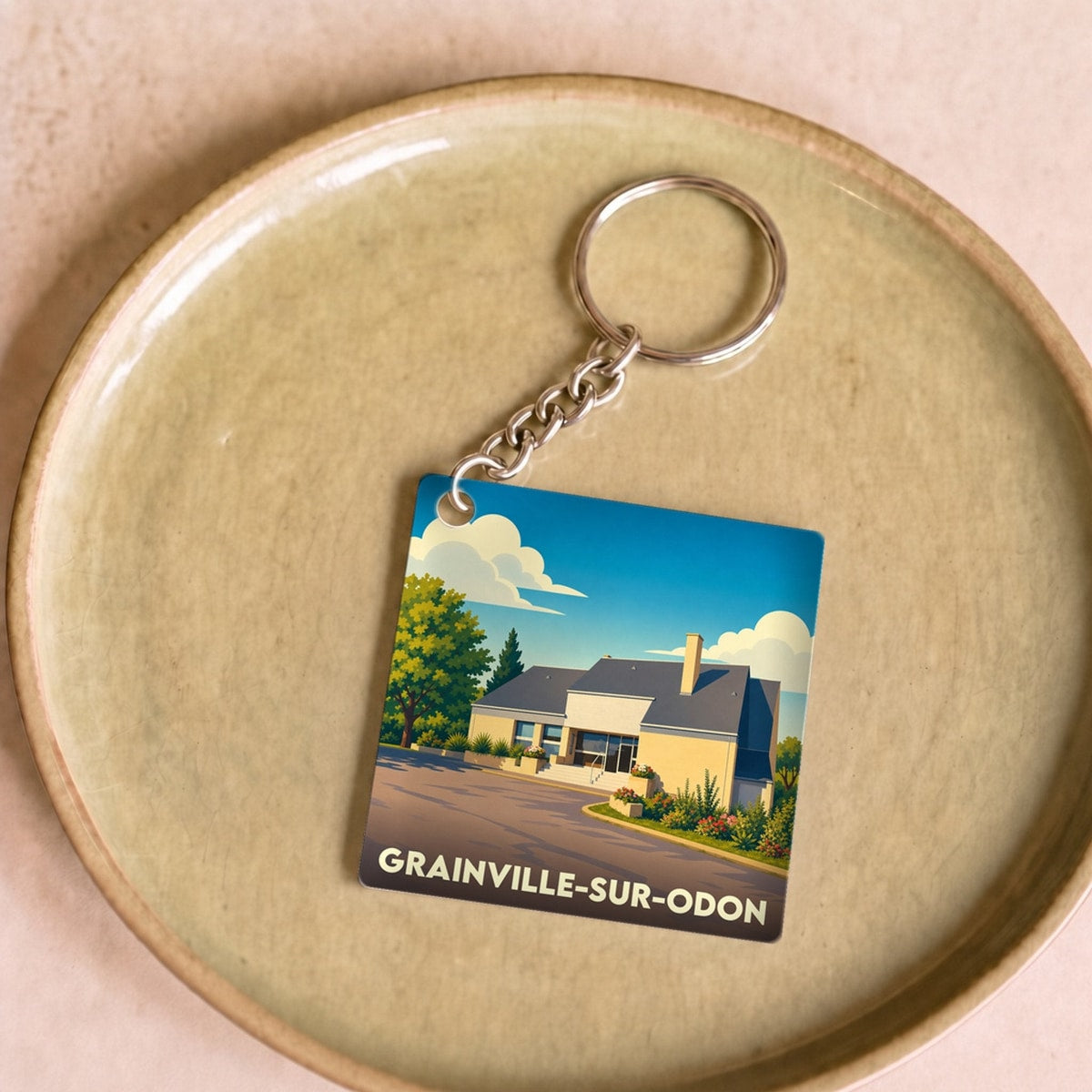 Porte-clés souvenir de Grainville-sur-Odon avec une illustration stylisée d'un bâtiment sous un ciel bleu.