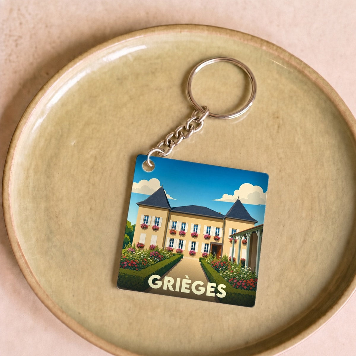 Porte-clés souvenir de la ville de Grièges avec une illustration stylisée d'une demeure bourgeoise fleurie.