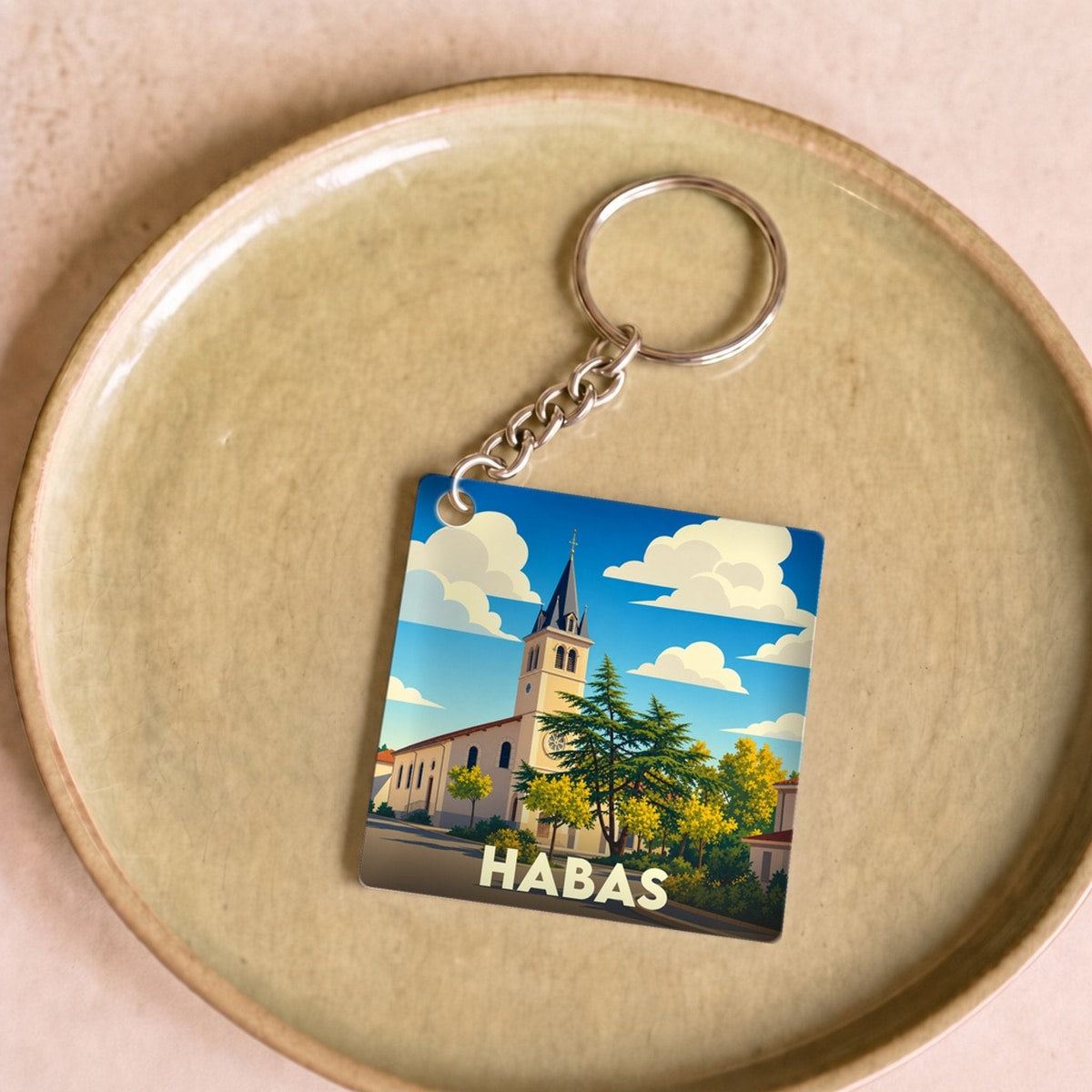Porte-clés souvenir de la ville de Habas avec une illustration stylisée de son église sous un ciel nuageux.