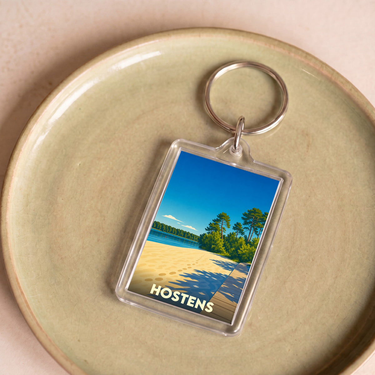 Porte-clés souvenir de la ville de Hostens avec une illustration stylisée d'une plage au bord d'un lac.