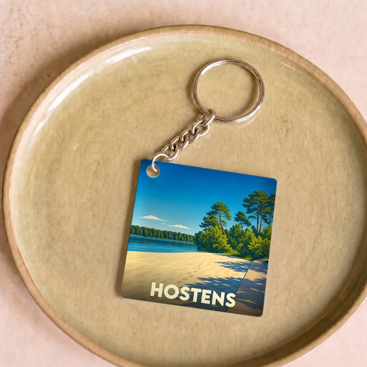 Porte-clés souvenir de la ville de Hostens avec une illustration stylisée d'une plage au bord d'un lac.