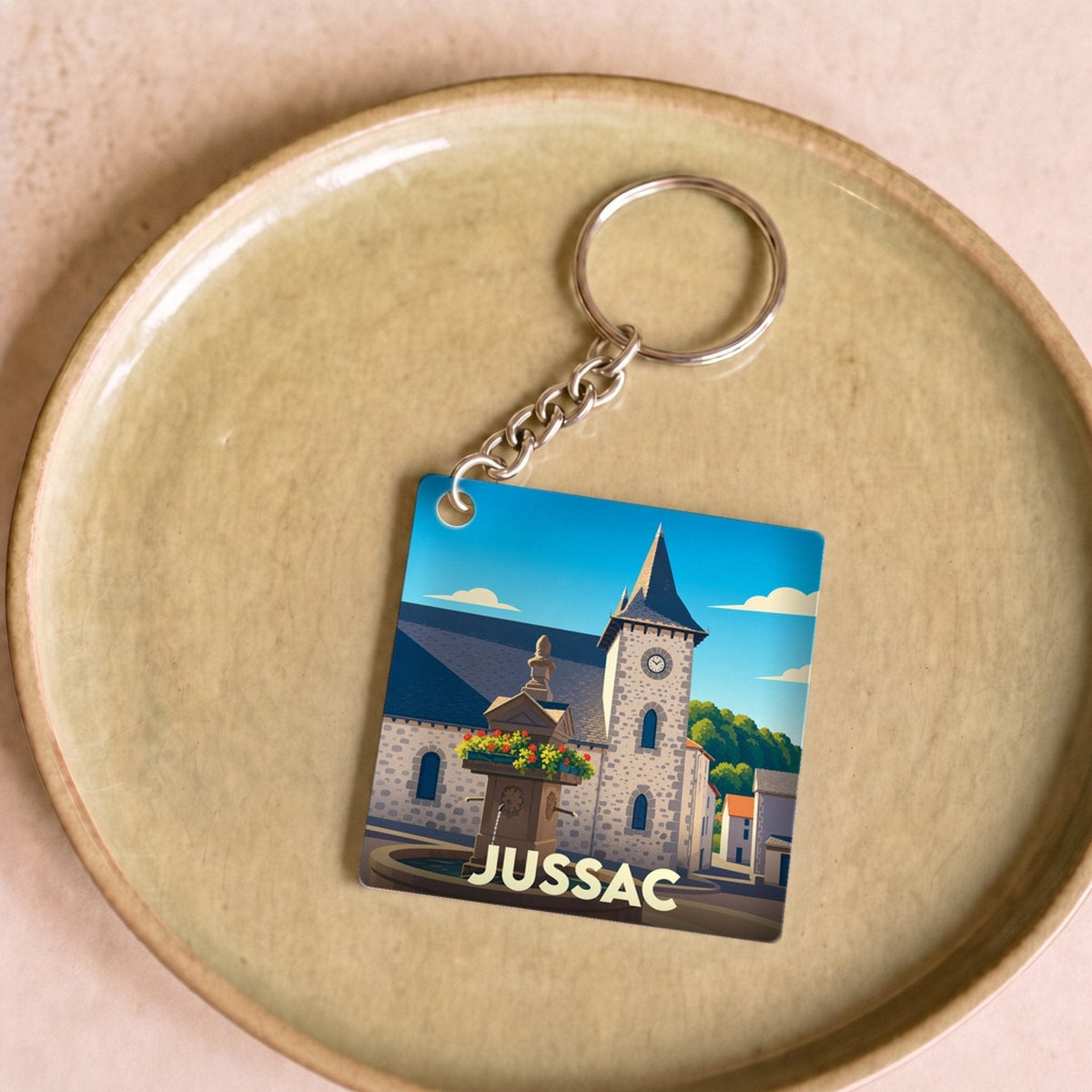 Porte-clés souvenir de la ville de Jussac avec une illustration stylisée d'une fontaine et de son église.