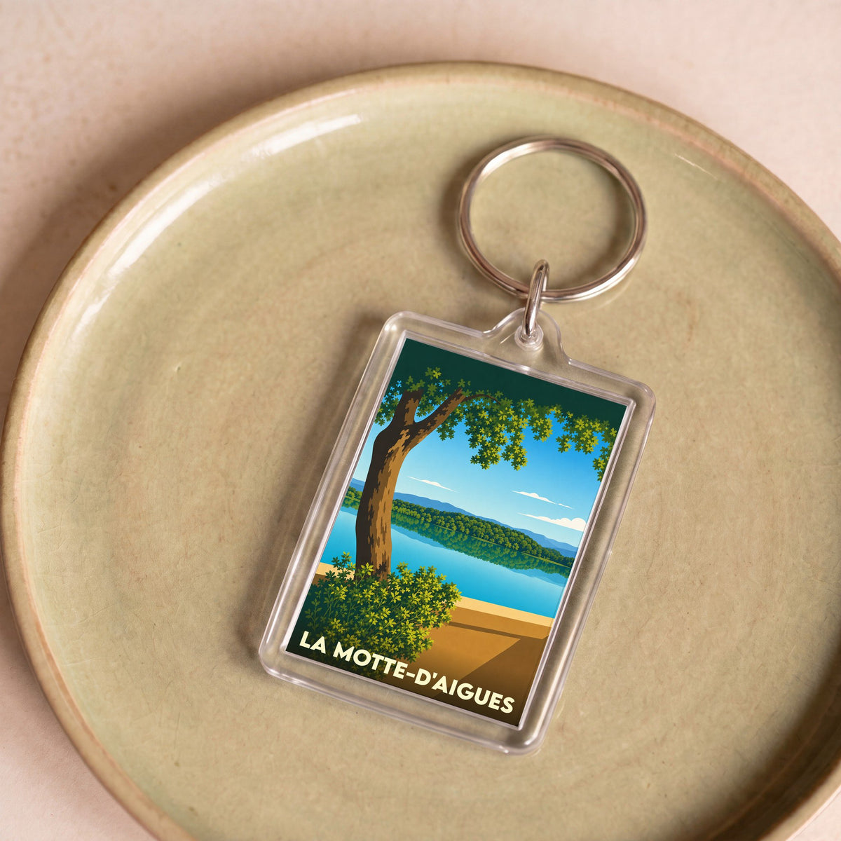Porte-clés souvenir de la ville de La Motte-d'Aigues avec une illustration stylisée d'un lac bordé d'arbres.