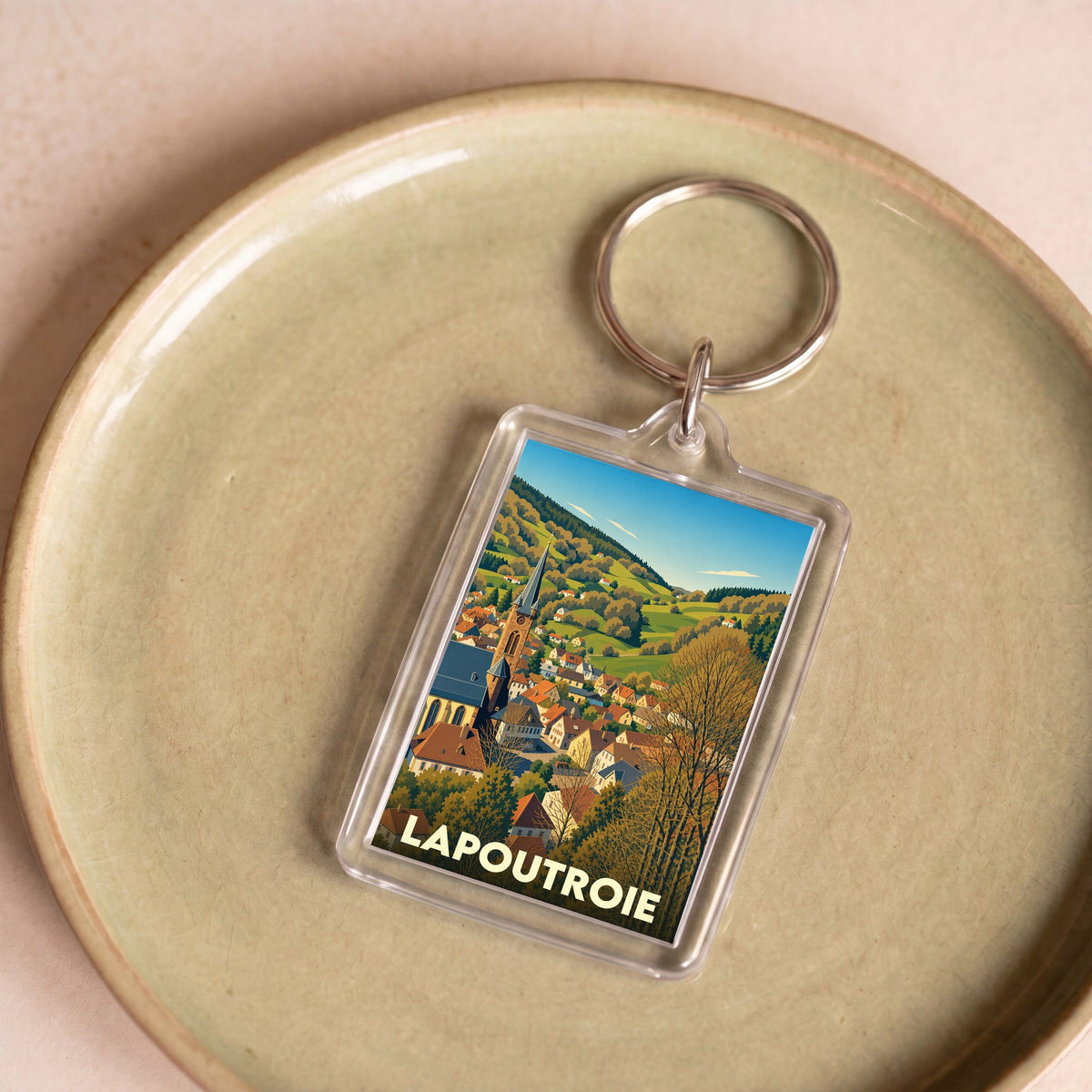 Porte-clés souvenir de la ville de Lapoutroie avec une illustration stylisée d'un village alsacien niché dans une vallée verdoyante.