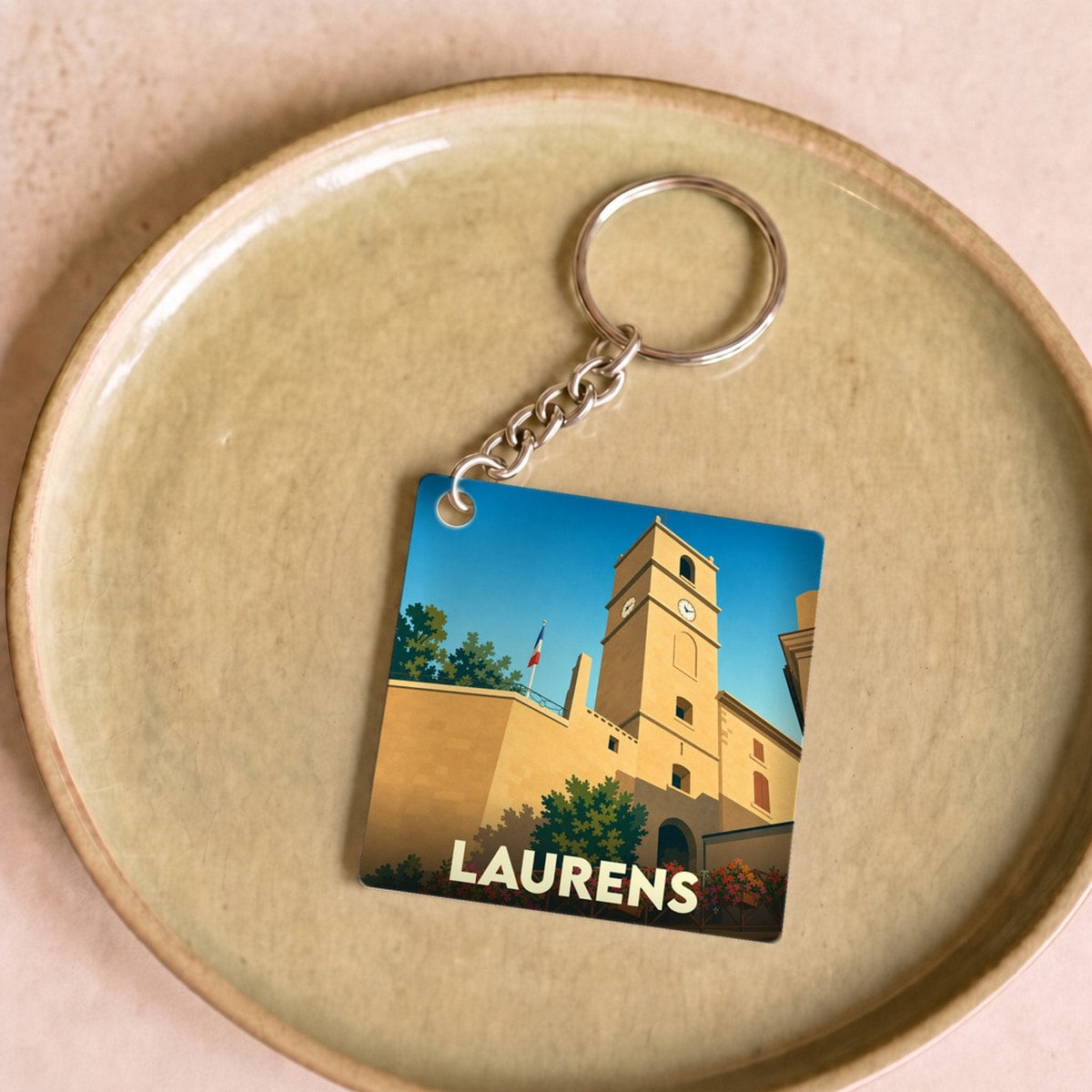 Porte-clés souvenir de la ville de Laurens avec une illustration stylisée de son clocher sous un ciel bleu.