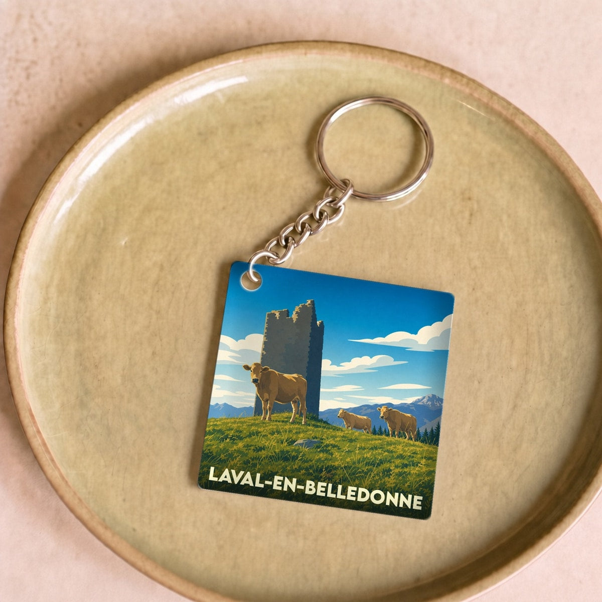 Porte-clés souvenir de Laval-en-Belledonne avec une illustration stylisée montrant des vaches devant une tour en pierre.