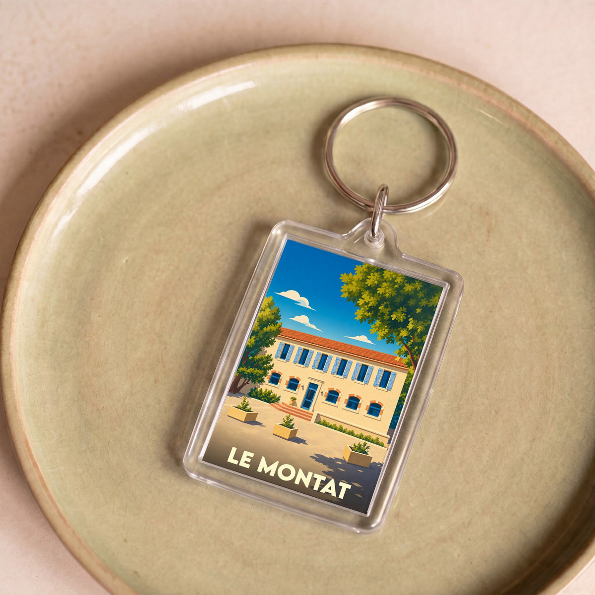Porte-clés souvenir de la ville de Le Montat avec une illustration stylisée d'une bâtisse ensoleillée.