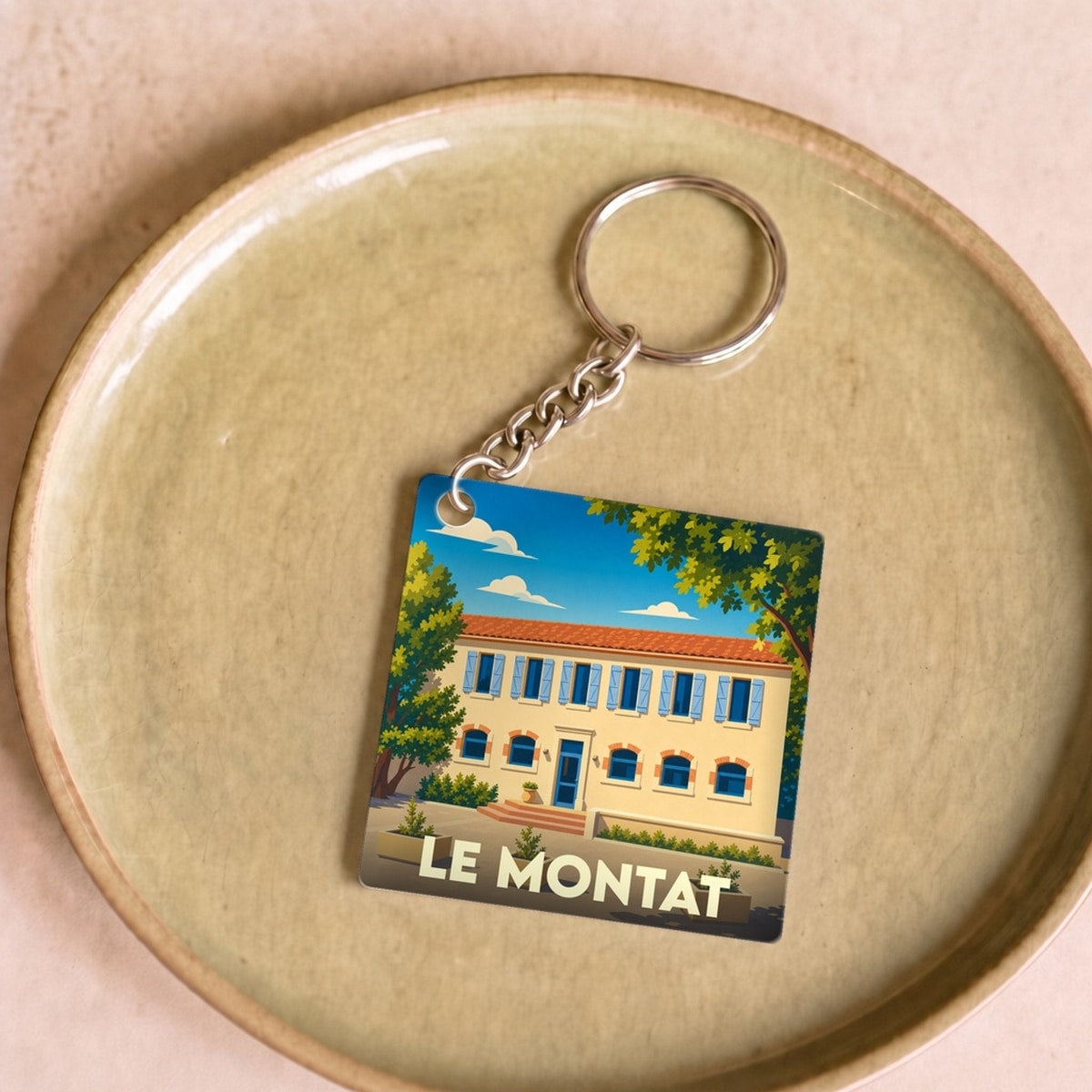 Porte-clés souvenir de la ville de Le Montat avec une illustration stylisée d'une bâtisse ensoleillée.
