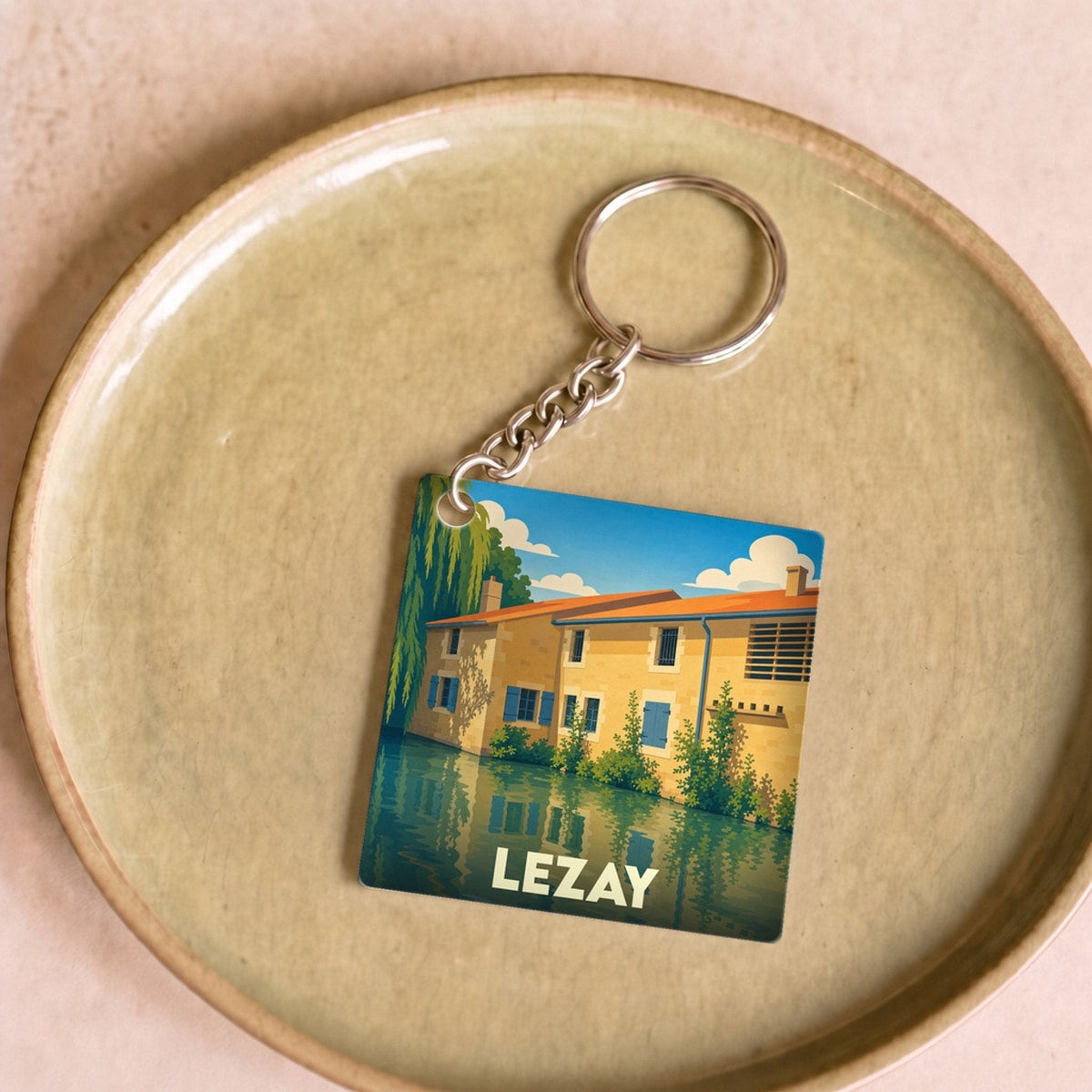 Porte-clés souvenir de la ville de Lezay avec une illustration colorée et stylisée de maisons au bord de l'eau.