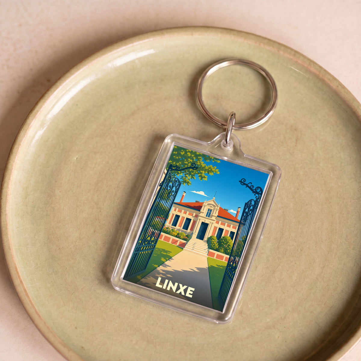 Porte-clés souvenir de la ville de Linxe avec une illustration stylisée d'une demeure bourgeoise derrière une grille.