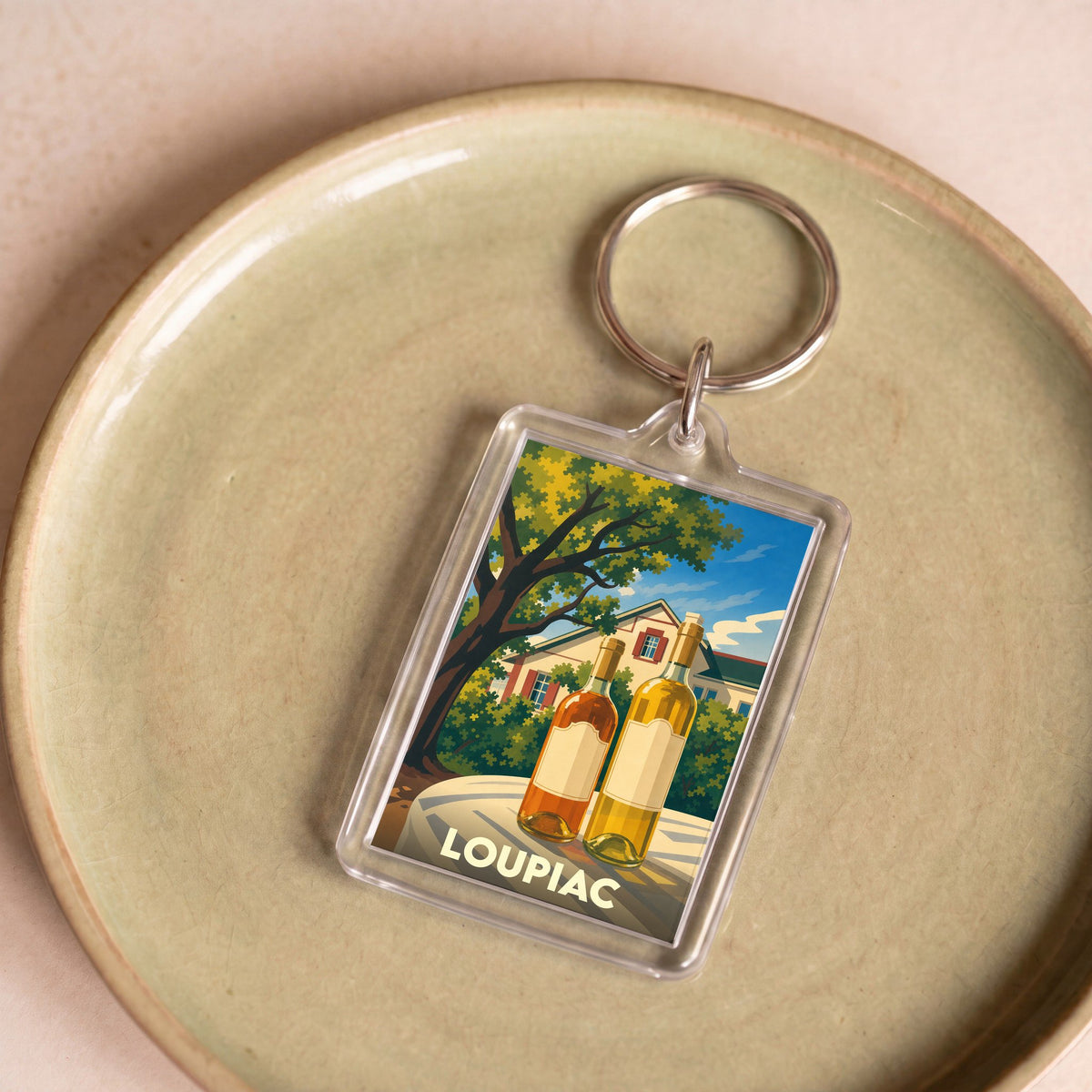 Porte-clés souvenir de la ville de Loupiac avec une illustration stylisée de bouteilles de vin devant une maison.