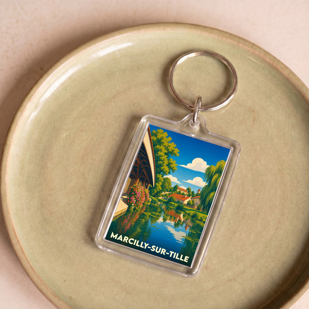 Porte-clés souvenir de Marcilly-sur-Tille avec une illustration colorée et stylisée de la ville au bord de l'eau.