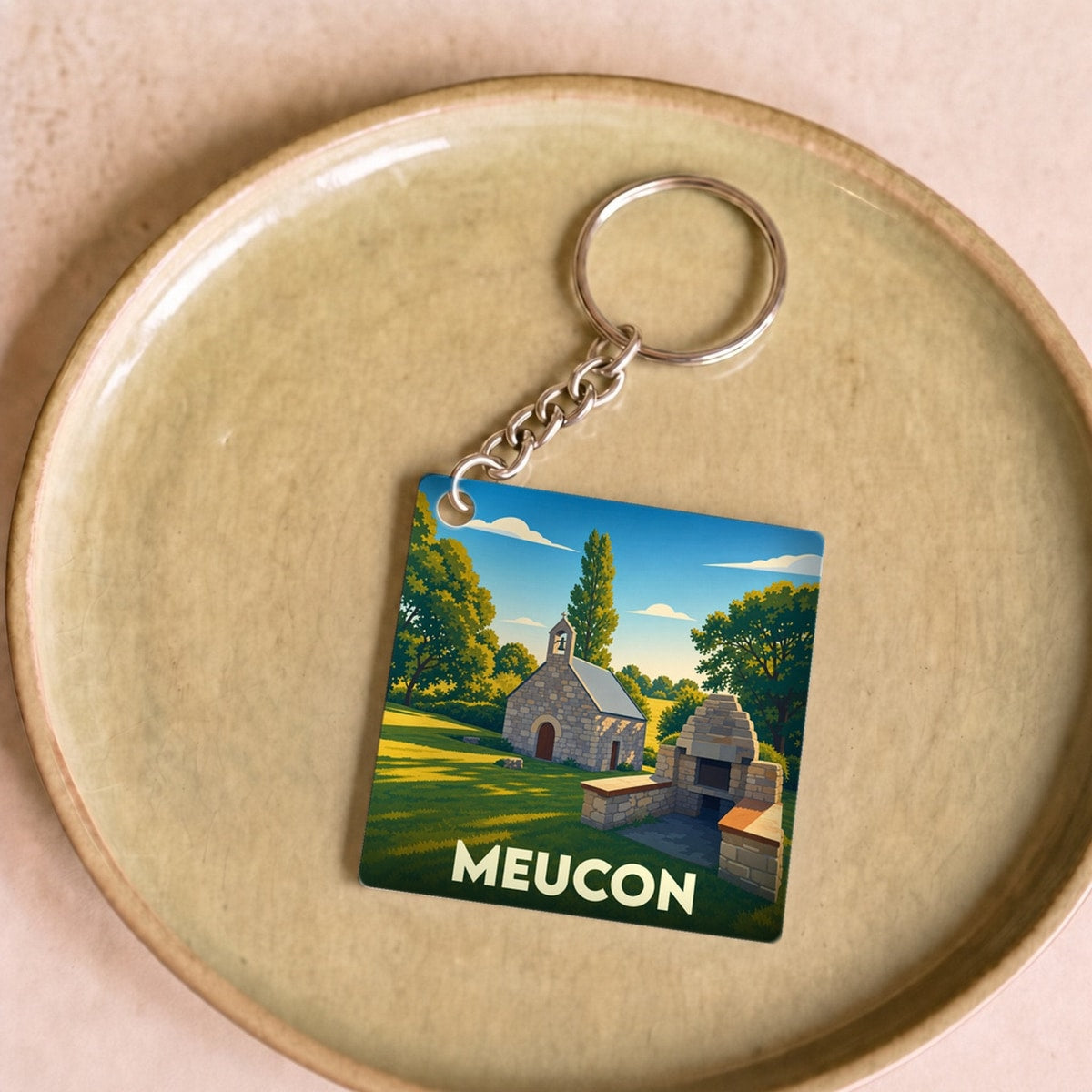 Porte-clés souvenir de la ville de Meucon avec une illustration stylisée d'une chapelle en pierre dans un paysage verdoyant.