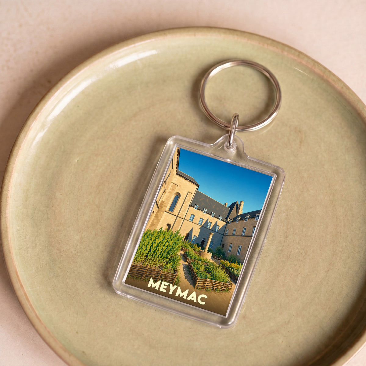 Porte-clés souvenir de la ville de Meymac avec une illustration stylisée de son abbaye et de ses jardins.