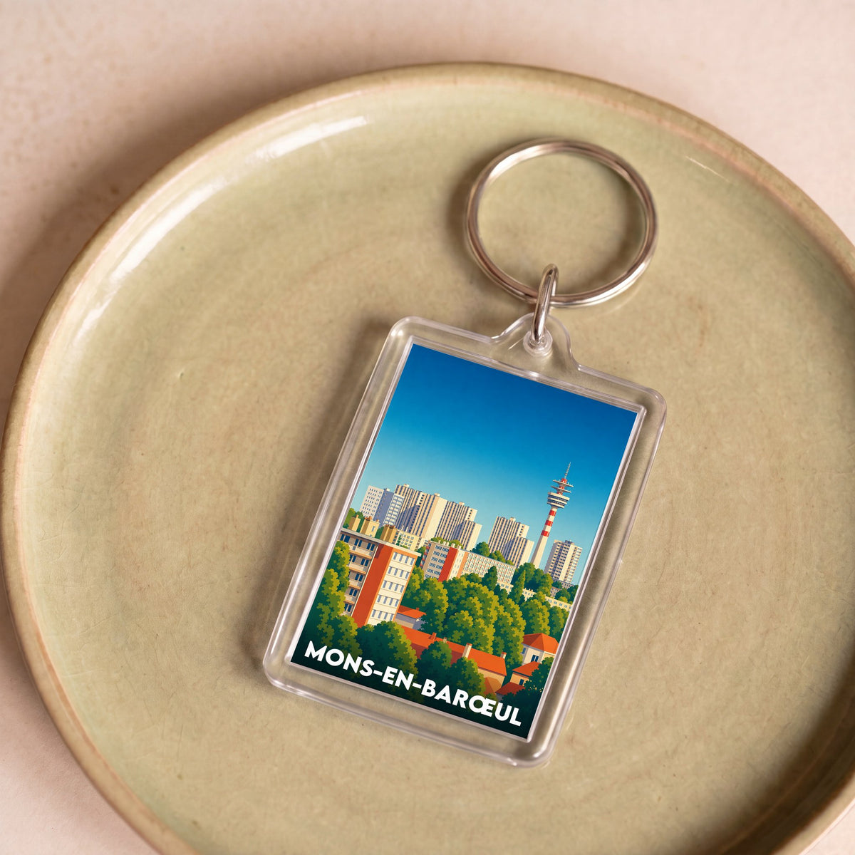 Porte-clés souvenir de la ville de Mons-en-Barœul avec une illustration graphique stylisée de son paysage urbain.
