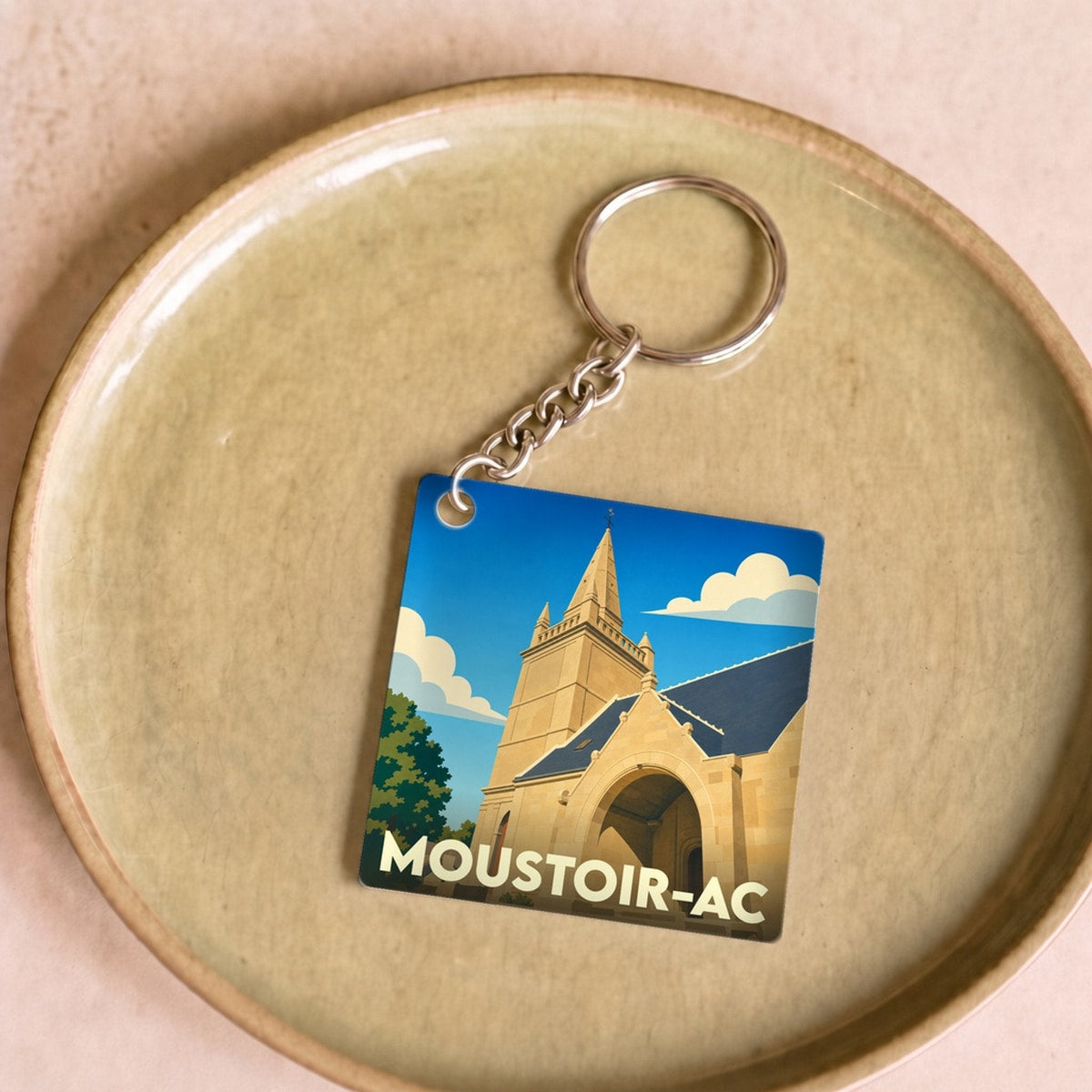 Porte-clés souvenir de la ville de Moustoir-Ac avec une illustration stylisée d'une église sous un ciel bleu.