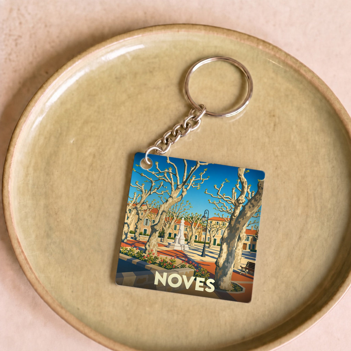 Porte-clés souvenir de la ville de Noves avec une illustration stylisée d'une place ensoleillée.