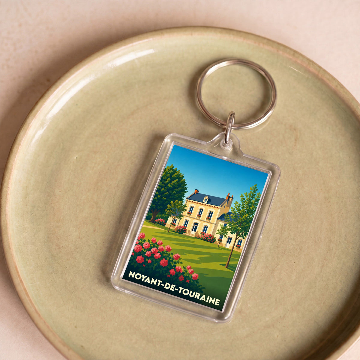 Porte-clés souvenir de la ville de Noyant-de-Touraine avec une illustration stylisée d'une maison bourgeoise et de son jardin.