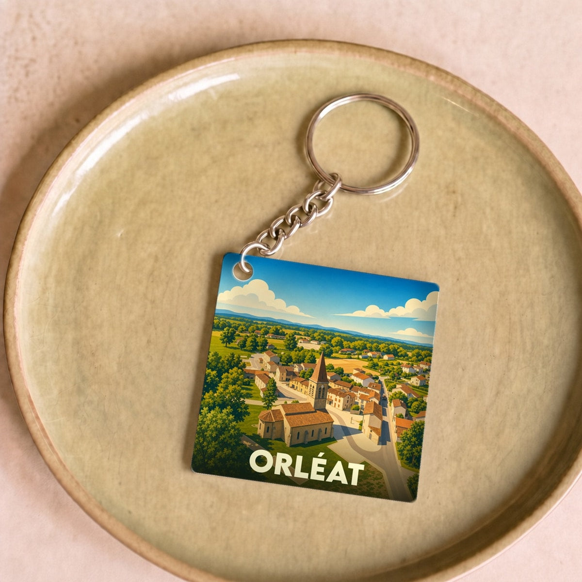 Porte-clés souvenir de la ville d'Orléat avec une illustration stylisée d'un paysage de village.